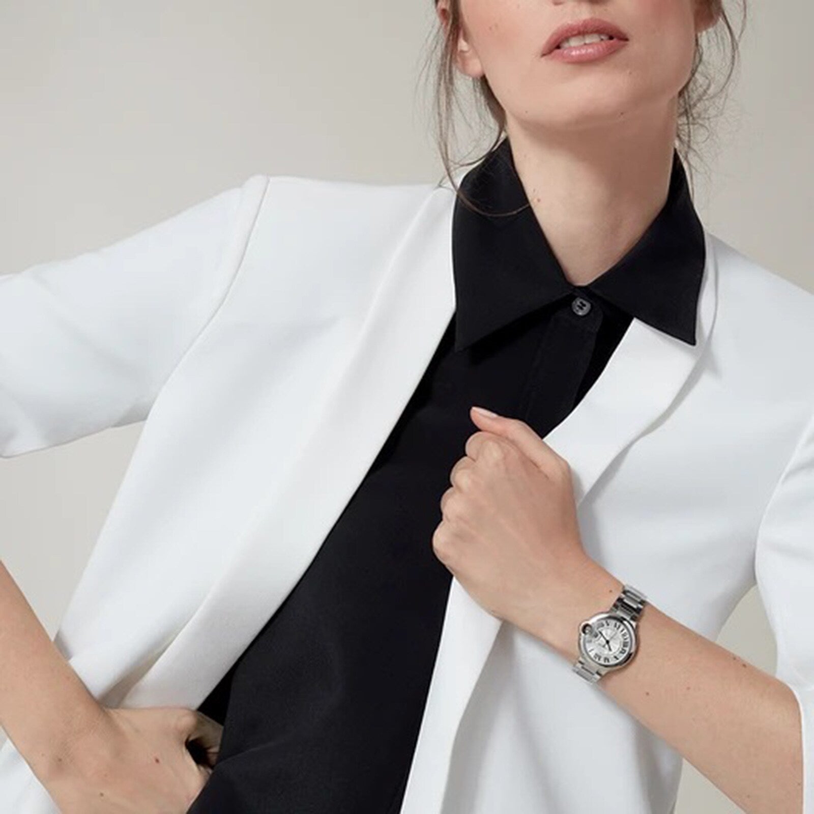 A woman in a white blazer over a black shirt, hand on her chest, wears a Cartier Ballon Bleu de Cartier watch with a silver guilloch dial on her left wrist.
