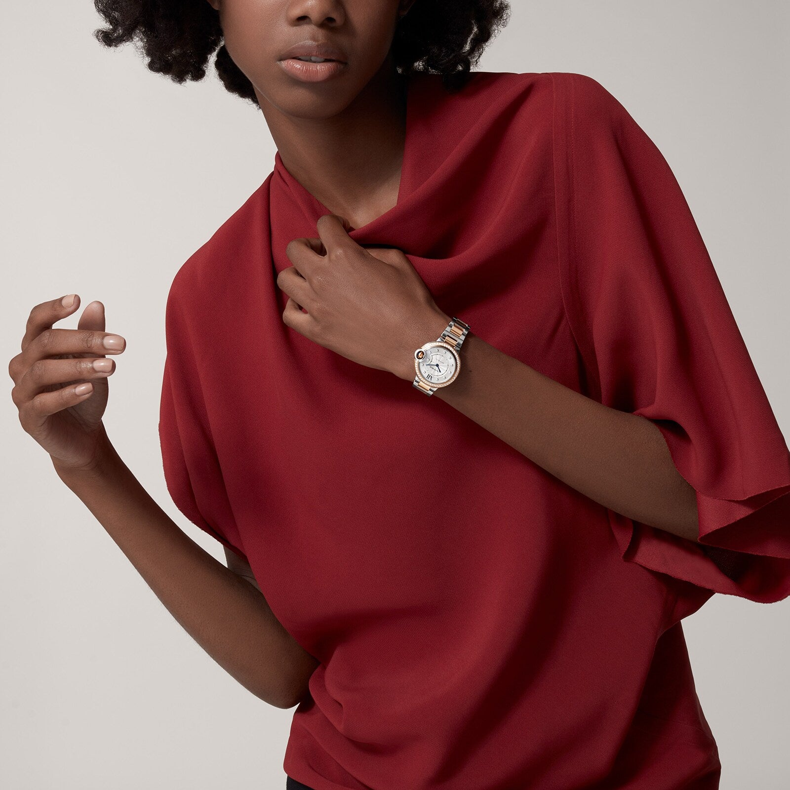 A person wearing a loose-fitting, short-sleeved red top showcases a sparkling Ballon Bleu de Cartier watch by Cartier with one hand near the collar.