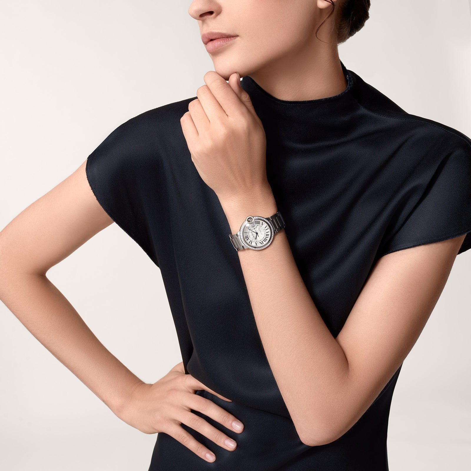 A woman in a black short-sleeve dress models the Cartier Ballon Bleu de Cartier silver mechanical wristwatch, one hand on her hip and the other near her face against a plain background.
