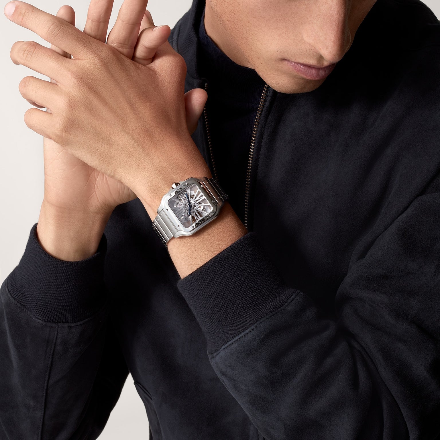 A person wearing a dark zip-up jacket showcases a Cartier Santos de Cartier Skeleton watch with mechanical movement and a square face on their left wrist.