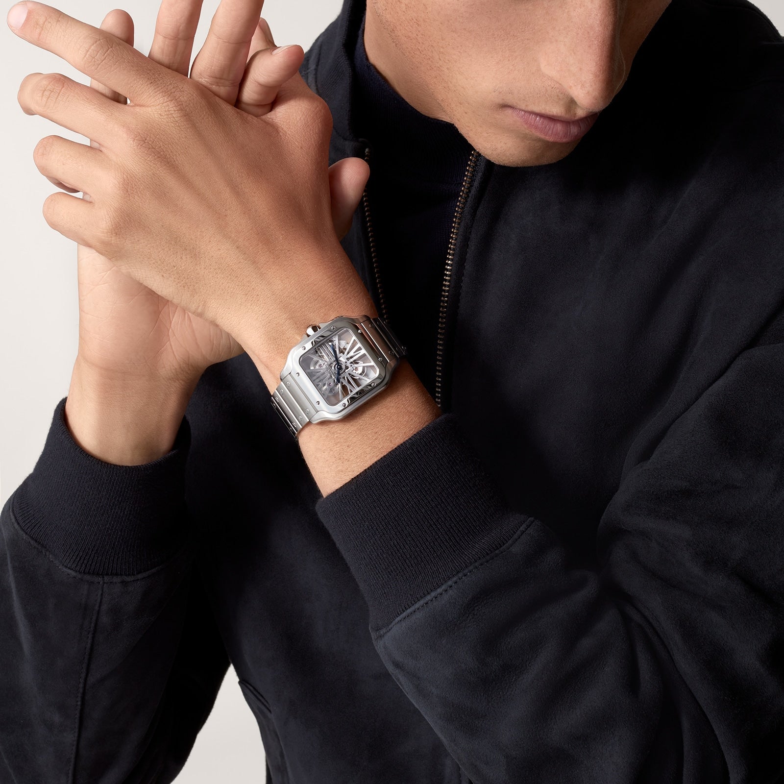A person wearing a dark zip-up jacket showcases a Cartier Santos de Cartier Skeleton watch with mechanical movement and a square face on their left wrist.