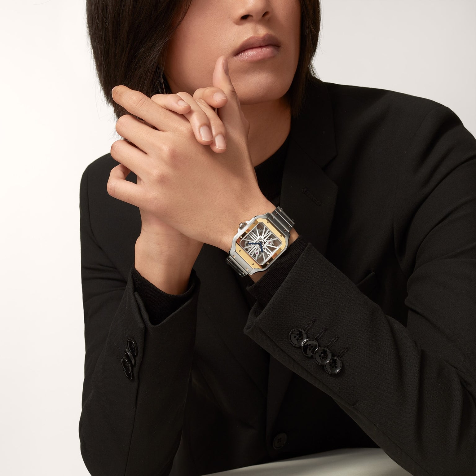 A person in a black suit with long hair rests their hands, showcasing the Cartier Santos de Cartier Skeleton watch with a silver and gold case, black strap, rectangular face, and visible mechanical movement.