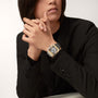 A person in a black suit with long hair rests their hands, showcasing the Cartier Santos de Cartier Skeleton watch with a silver and gold case, black strap, rectangular face, and visible mechanical movement.