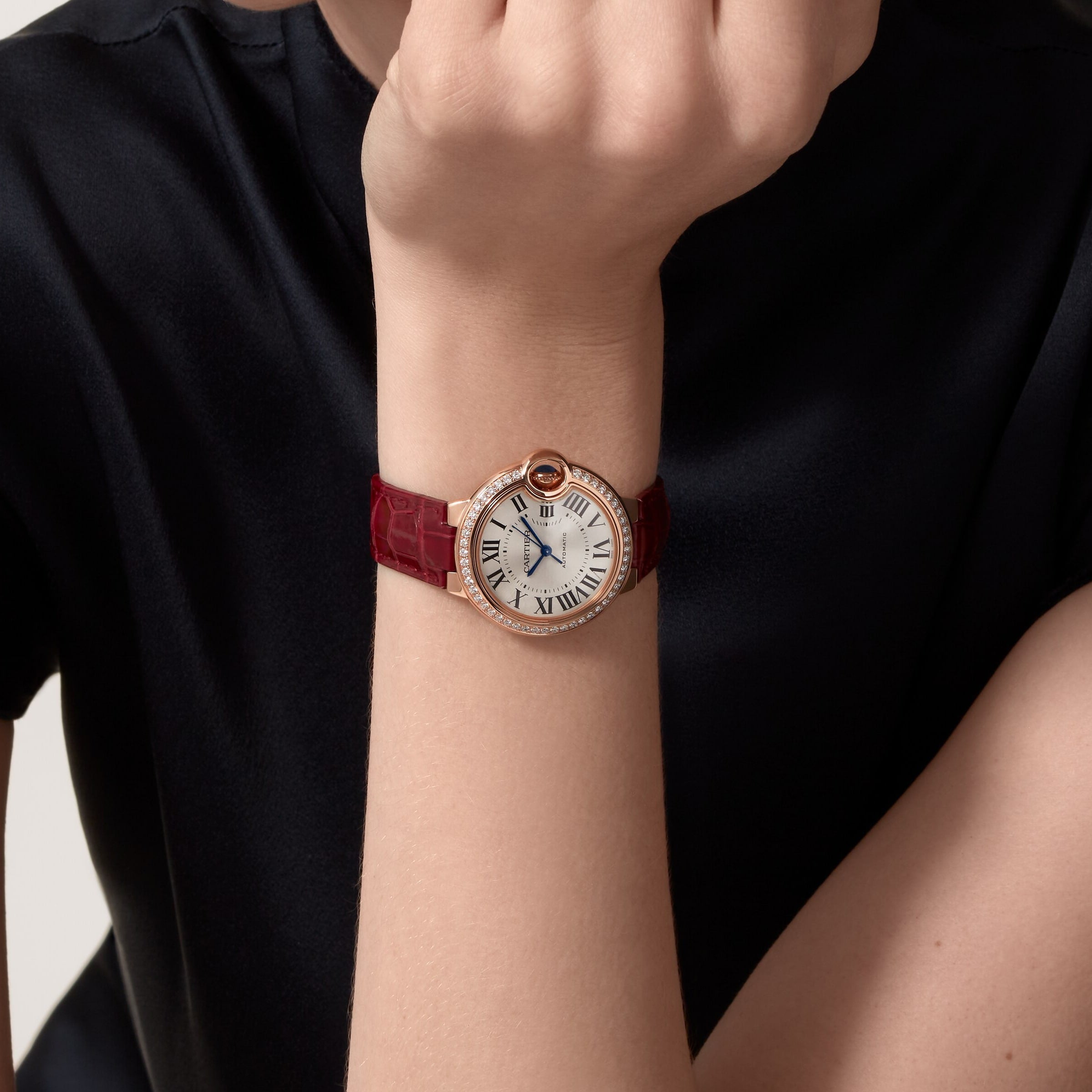 A person in a black shirt displays the Ballon Bleu de Cartier watch with a white face, Roman numerals, rose gold accents, diamond details, a red bezel, and a burgundy strap from Cartier.
