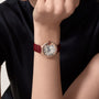 A person in a black shirt displays the Ballon Bleu de Cartier watch with a white face, Roman numerals, rose gold accents, diamond details, a red bezel, and a burgundy strap from Cartier.
