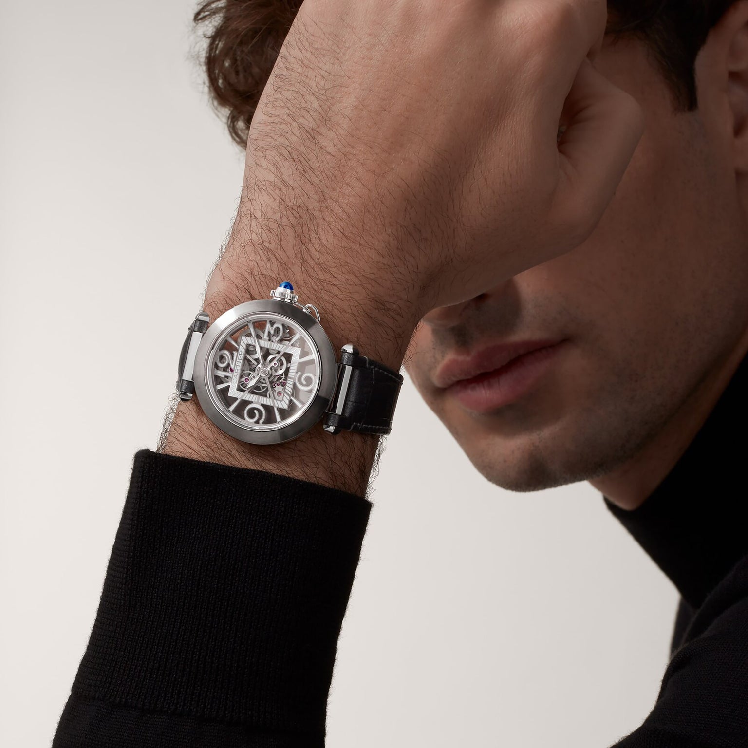 A man in a black long-sleeve shirt showcases the Cartier Pasha de Cartier Skeleton watch, featuring a striking skeleton dial and black band, his raised arm partly covering his face to highlight its impressive mechanical movement.
