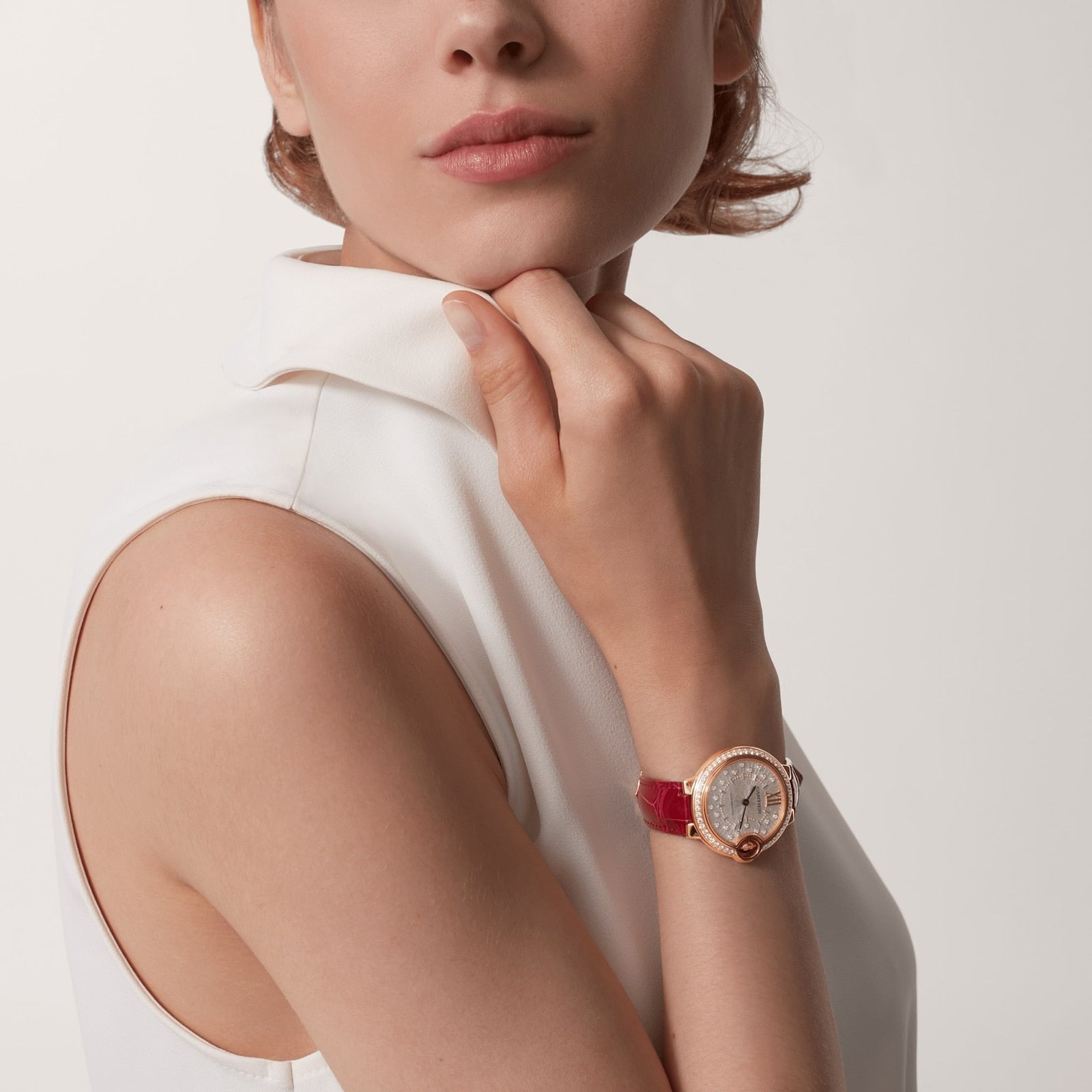 A woman in a sleeveless white top poses with her hand on her neck, wearing the Ballon Bleu de Cartier watch by Cartier with a red band on her wrist.