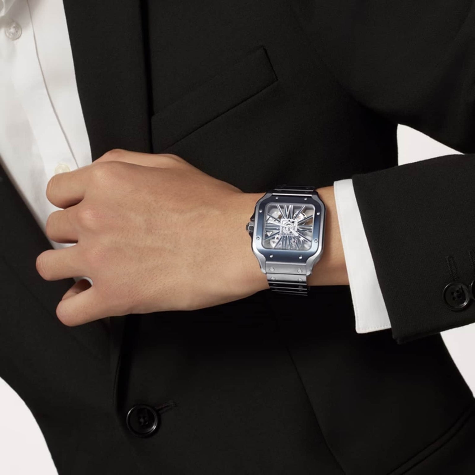 A person in a black suit and white shirt showcases a Cartier Santos de Cartier Skeleton watch with a skeleton design and Manufacture mechanical movement on their left wrist.