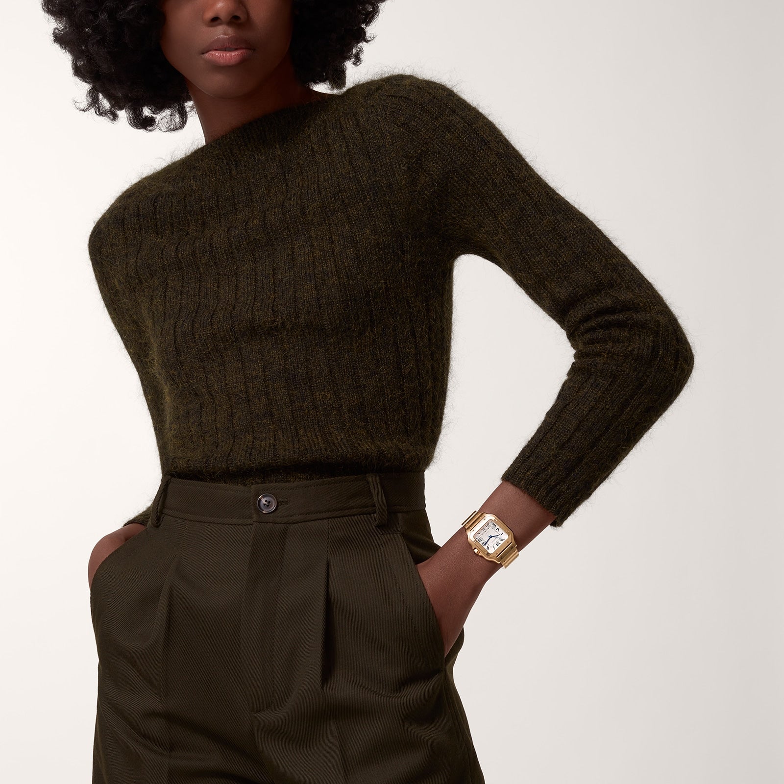 A person wearing a dark green knit sweater tucked into high-waisted, pleated dark green trousers, with a Cartier Baignoire watch in rose gold visible on their left wrist.