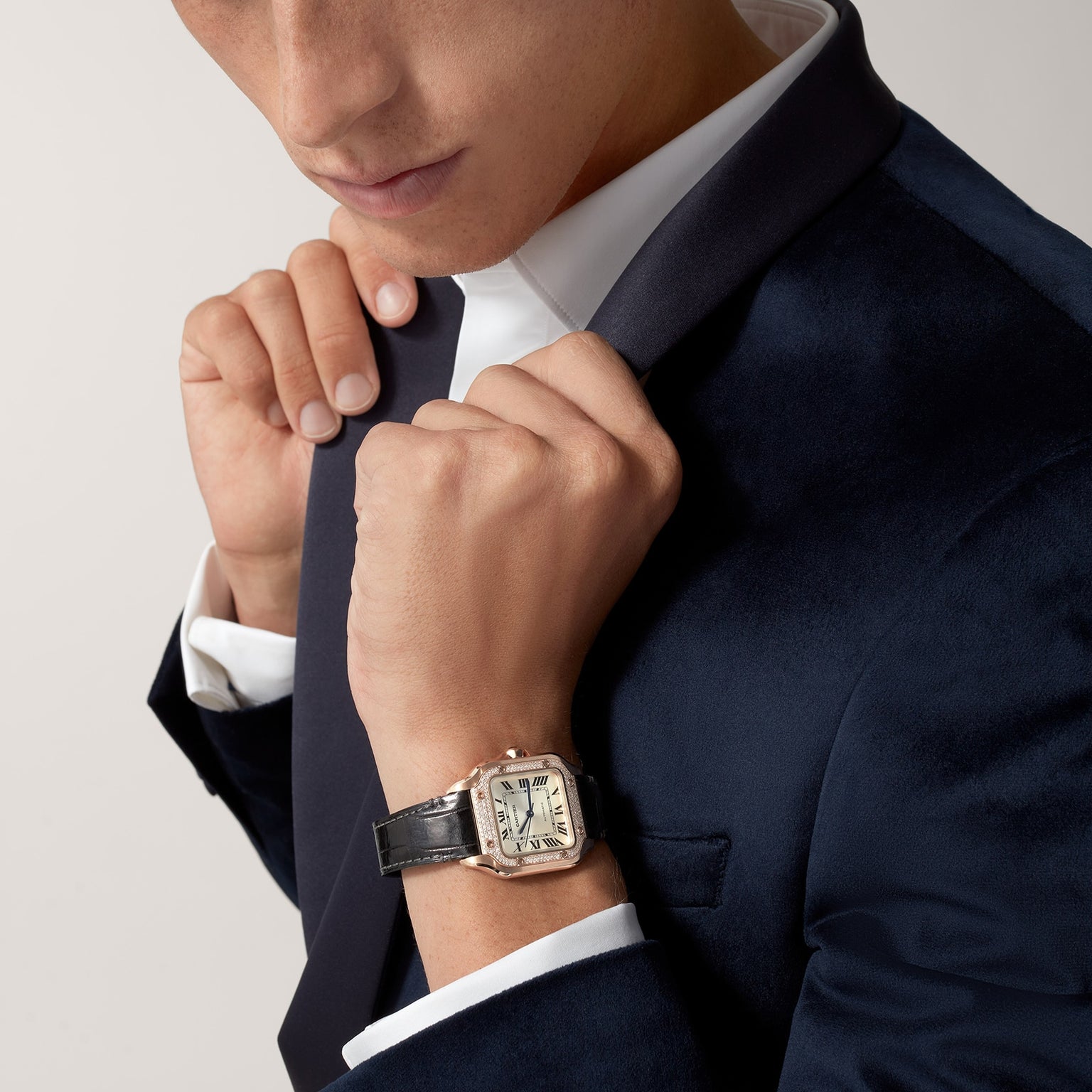 Man in a dark velvet suit and white shirt adjusts his collar, wearing a Santos de Cartier watch by Cartier with a rose gold case and black leather strap on his wrist.
