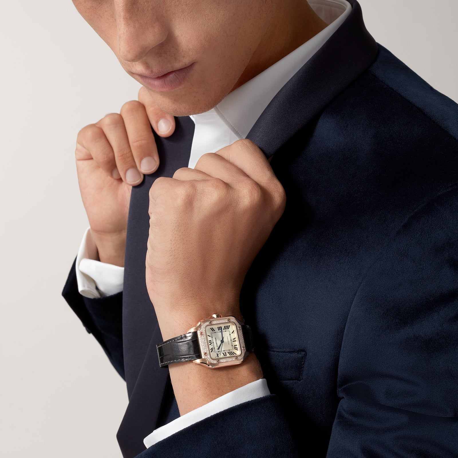 Man in a dark velvet suit and white shirt adjusts his collar, wearing a Santos de Cartier watch by Cartier with a rose gold case and black leather strap on his wrist.