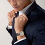 Man in a dark velvet suit and white shirt adjusts his collar, wearing a Santos de Cartier watch by Cartier with a rose gold case and black leather strap on his wrist.
