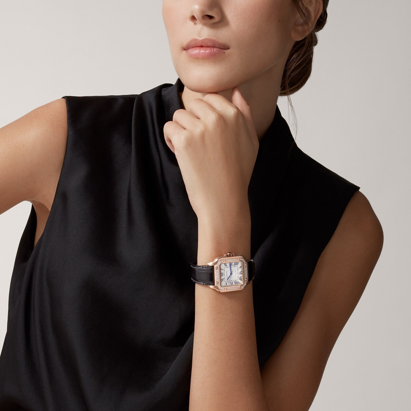 A woman in a sleeveless black top rests her chin on her hand, showcasing the Santos de Cartier watch by Cartier, featuring a square face, black strap, and rose gold accents.