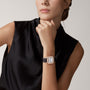 A woman in a sleeveless black top rests her chin on her hand, showcasing the Santos de Cartier watch by Cartier, featuring a square face, black strap, and rose gold accents.
