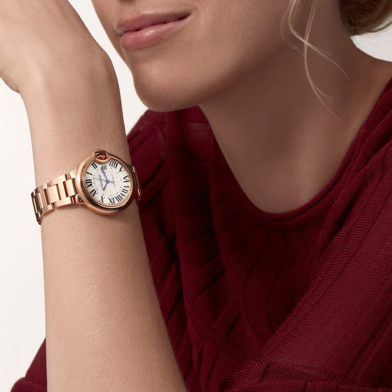 A person in a red sweater rests their chin on their hand, wearing a Cartier Ballon Bleu de Cartier wristwatch with a rose gold case, cabochon sapphire crown, and a white Roman numeral dial.