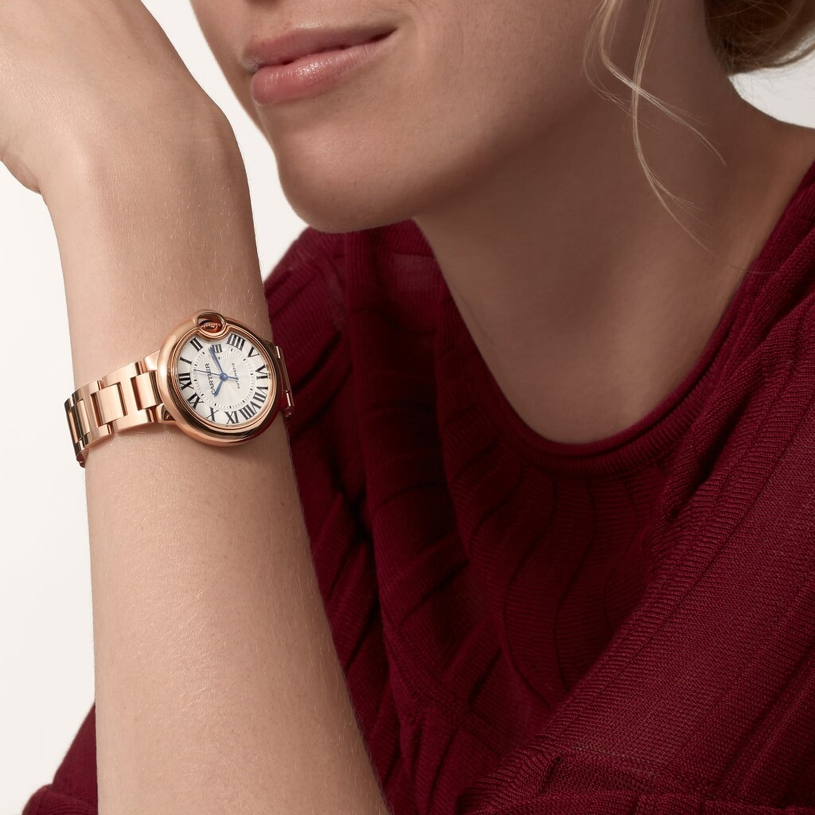 A person in a red sweater rests their chin on their hand, wearing a Cartier Ballon Bleu de Cartier wristwatch with a rose gold case, cabochon sapphire crown, and a white Roman numeral dial.
