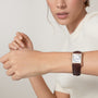 A woman in a white short-sleeve top showcases the Cartier Tank Louis Cartier watch, featuring a rectangular face with Roman numerals, brown leather strap, and signature sapphire cabochon crown.
