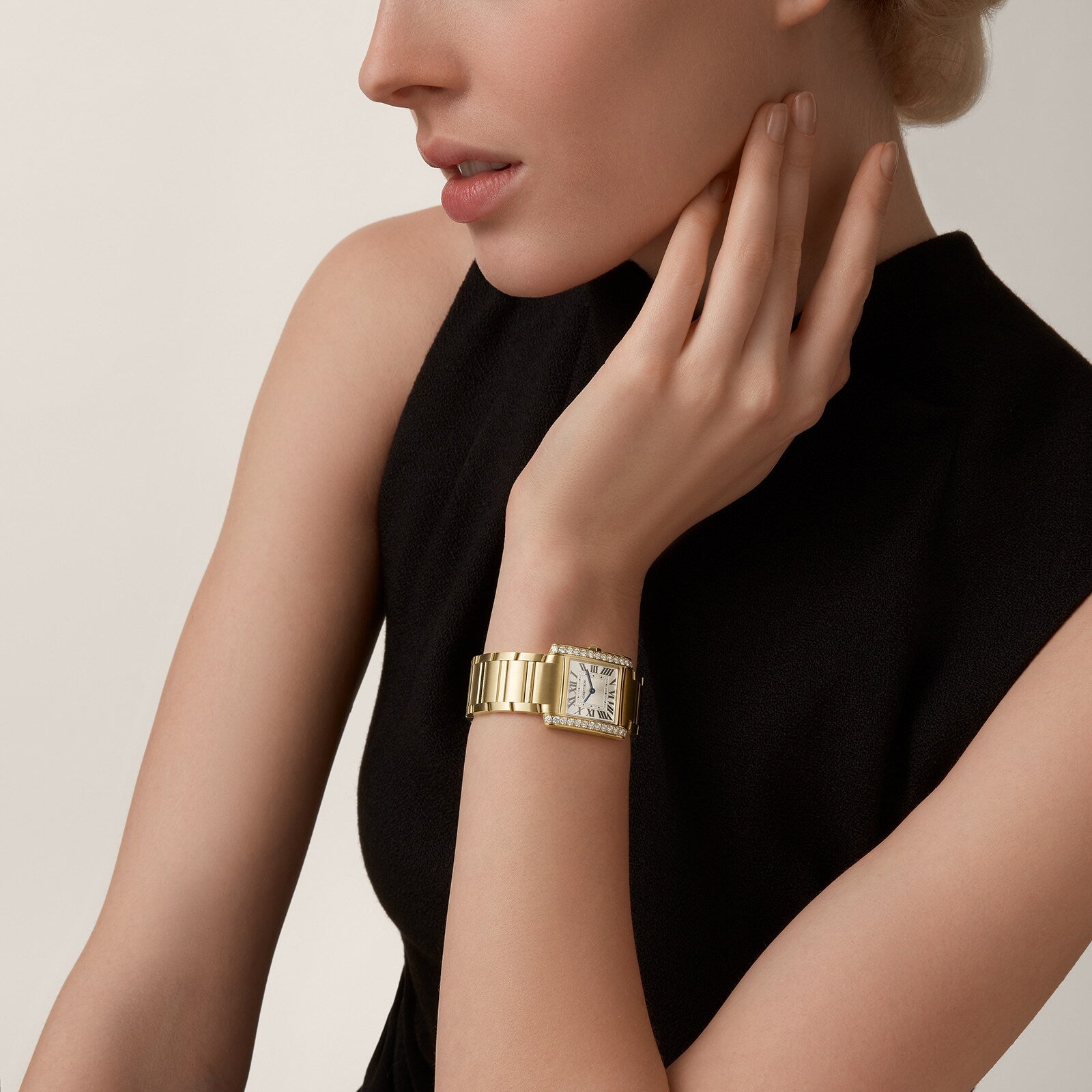 A woman in a sleeveless black dress rests her hand on her neck, wearing a Cartier Tank Américaine with a yellow gold case and brilliant-cut diamonds accenting its rectangular face.