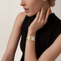 A woman in a sleeveless black dress rests her hand on her neck, wearing a Cartier Tank Américaine with a yellow gold case and brilliant-cut diamonds accenting its rectangular face.