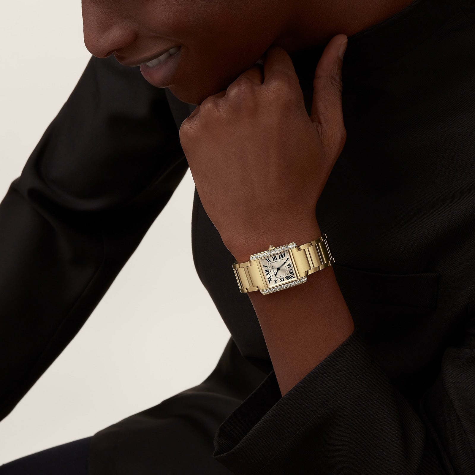 A person in a black top rests their chin on their hand, showcasing a Cartier Tank Américaine with a yellow gold case and brilliant-cut diamonds on the rectangular face.