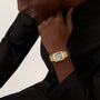 A person in a black top rests their chin on their hand, showcasing a Cartier Tank Américaine with a yellow gold case and brilliant-cut diamonds on the rectangular face.