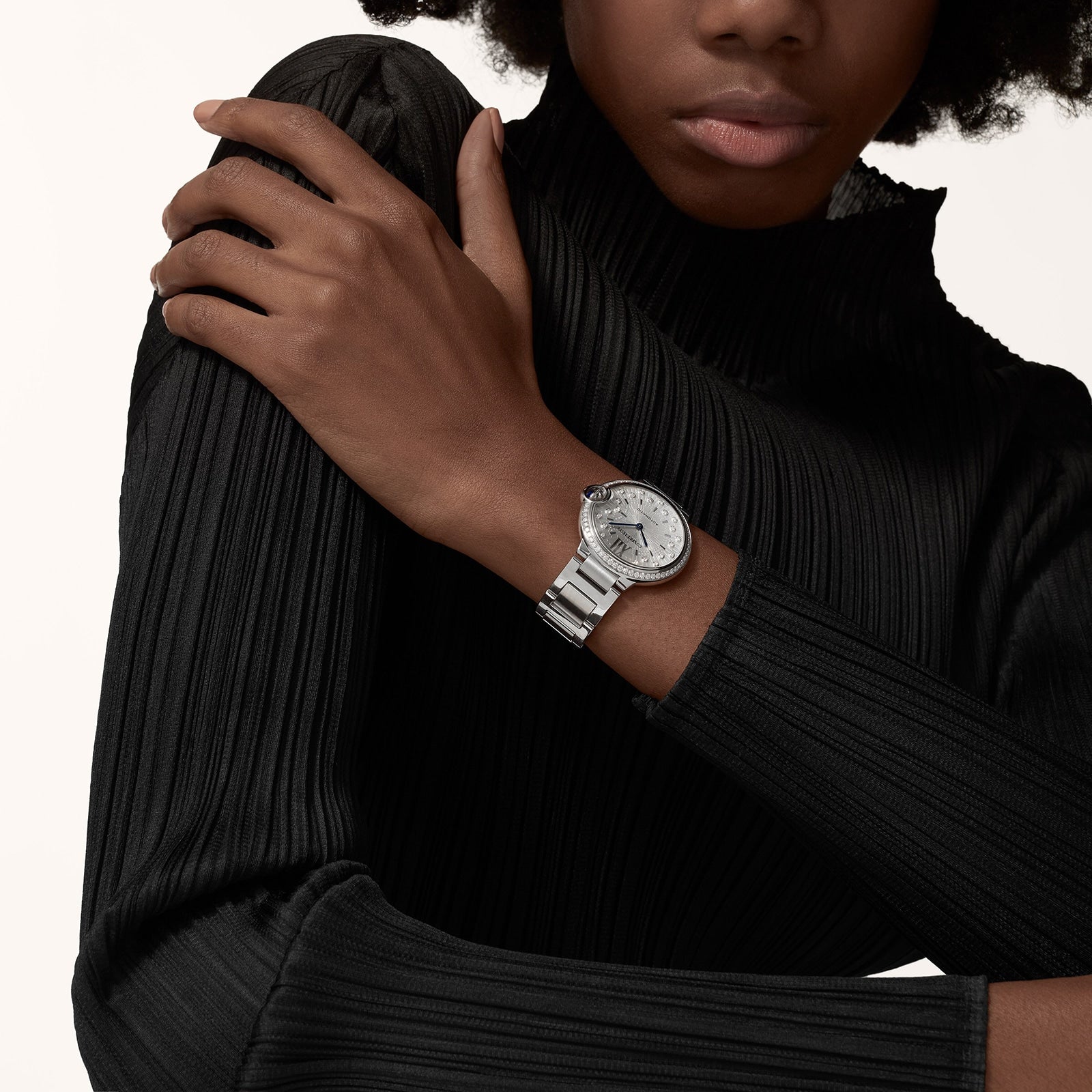 A person in a black, long-sleeved top poses with arms crossed, displaying a Cartier Ballon Bleu de Cartier with an interchangeable steel bracelet and sunray grey dial on their left wrist.
