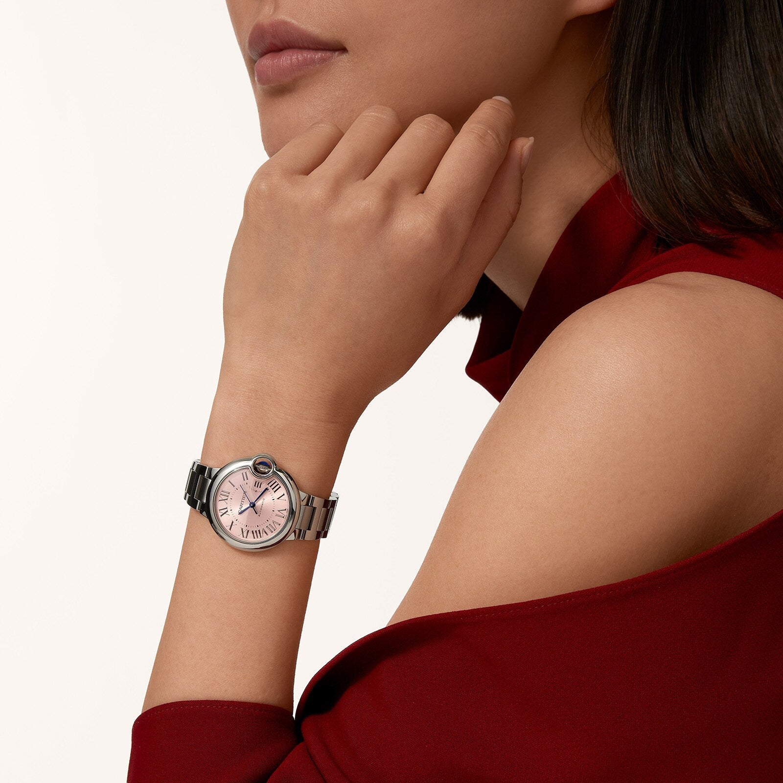 A woman in a burgundy top rests her chin on her hand, showcasing the Cartier Ballon Bleu wristwatch with elegant Roman numeral markers and a pink sunray dial.