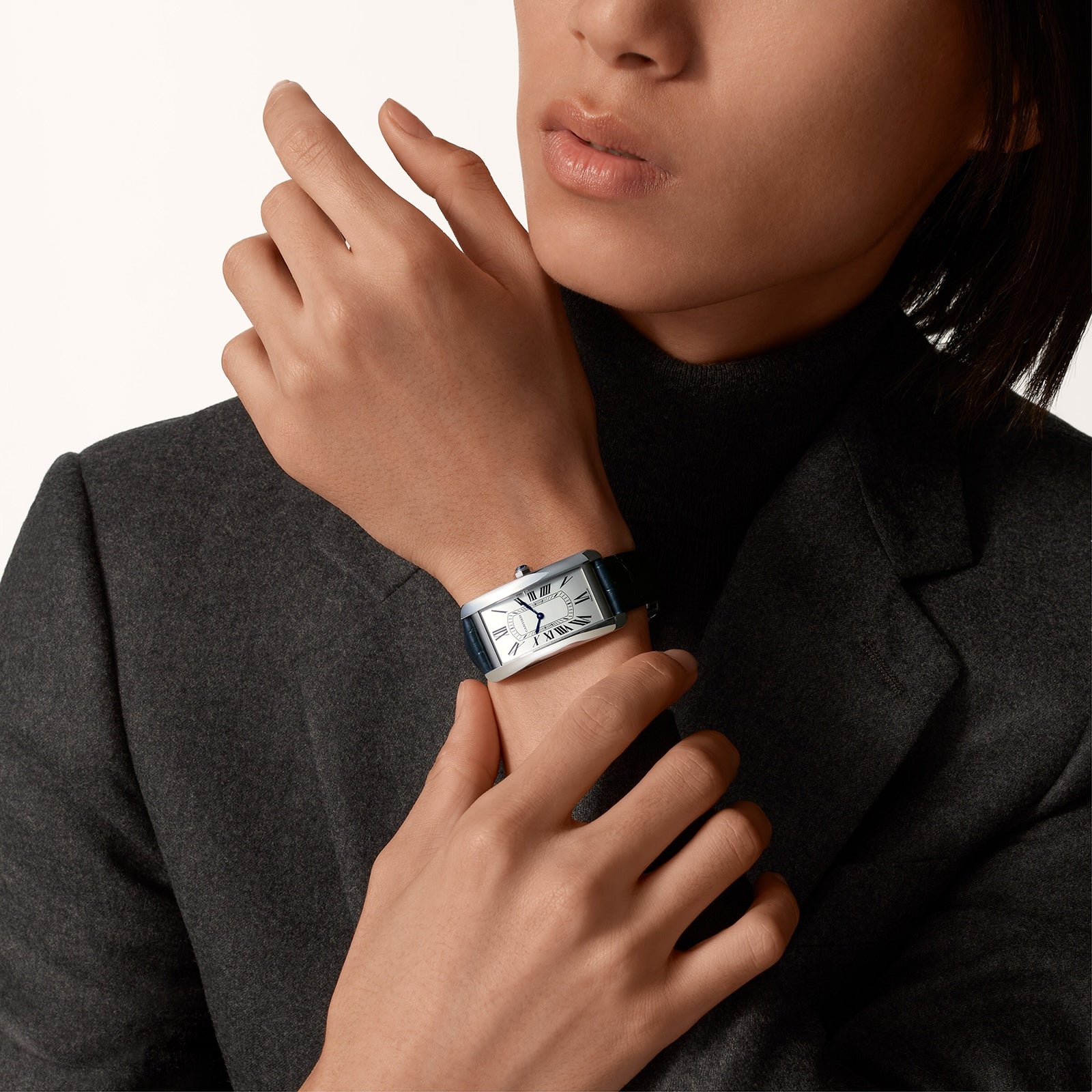 Wearing a dark blazer and turtleneck, a person showcases the Cartier Tank Américaine—a sleek rectangular silver watch with a black strap—on their left wrist, embodying timeless elegance.