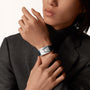 Wearing a dark blazer and turtleneck, a person showcases the Cartier Tank Américaine—a sleek rectangular silver watch with a black strap—on their left wrist, embodying timeless elegance.
