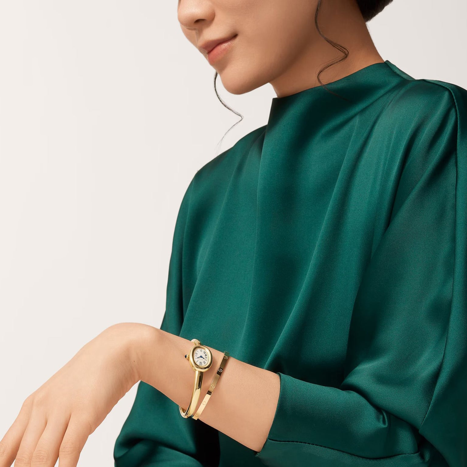 A woman in a green satin top smiles slightly, wearing a Cartier Baignoire (Size 17) gold wristwatch with a matching bracelet.