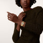 A person in a dark ribbed sweater showcases the Cartier Tank Française, featuring Roman numeral markers and a yellow gold bracelet, worn on their left wrist against a simple backdrop.