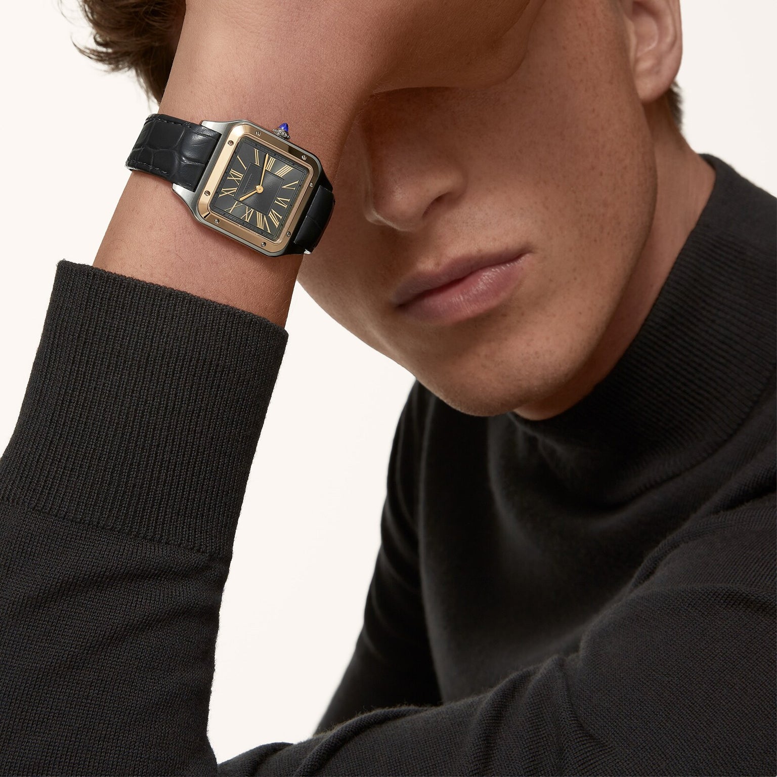 A person in a black turtleneck rests their head on their arm, showcasing the Cartier Santos-Dumont watch with an 18K yellow gold bezel, black leather strap, and Roman numerals—a true icon of Cartier design.
