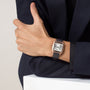 A person in a dark suit showcases the Cartier Santos-Dumont watch, featuring a white dial with black Roman numerals, a luxurious 18K rose gold case, and an elegant alligator leather strap.