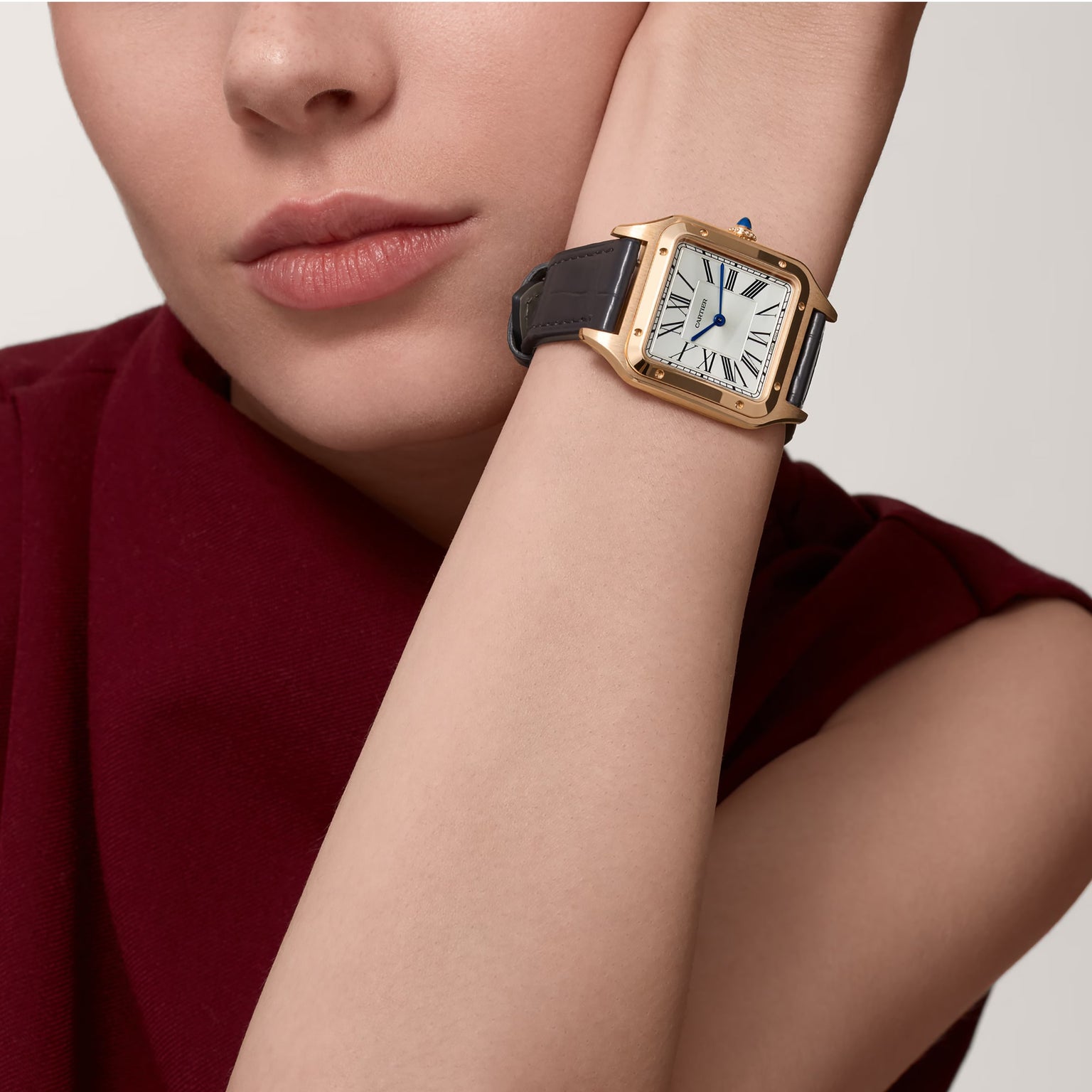 A person in a burgundy top, resting their chin on their hand, wears a Cartier Santos-Dumont watch featuring 18K rose gold, an alligator leather strap, and a Roman numeral dial.