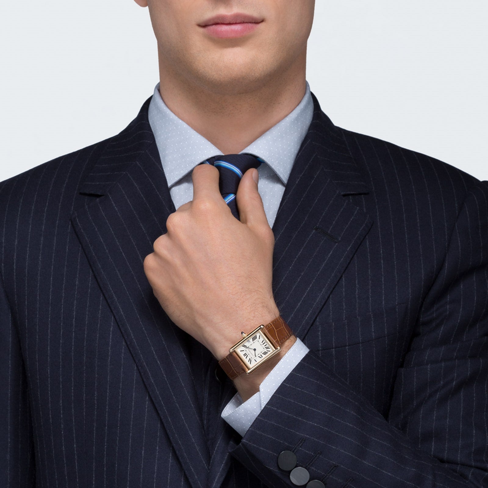 A man in a pinstripe suit adjusts his tie, wearing a Cartier Tank Louis Cartier watch in 18K yellow gold with a brown leather strap.
