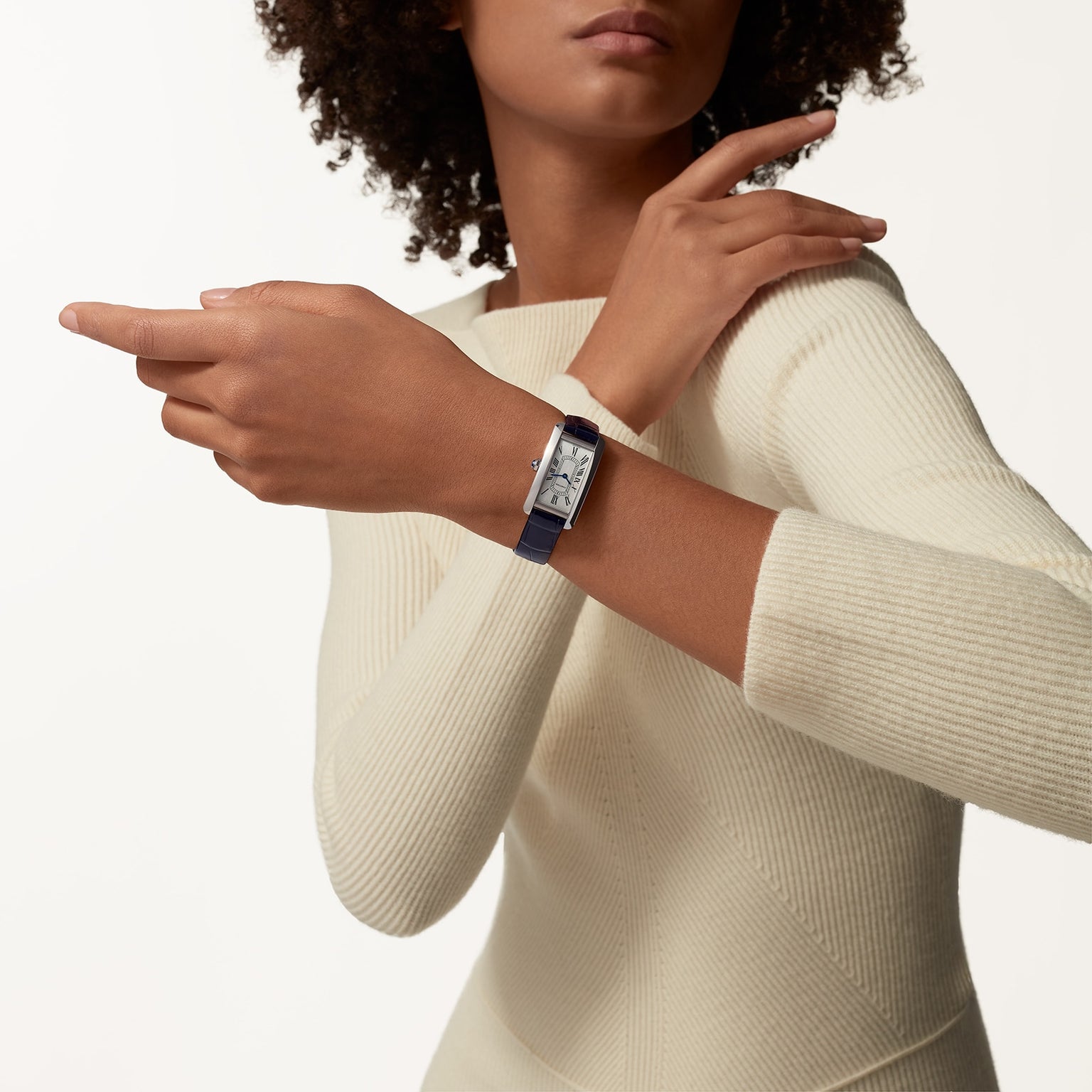 Against a plain background, a person in a cream ribbed sweater showcases the timeless elegance of the Cartier Tank Américaine watch with a dark strap on their left wrist.