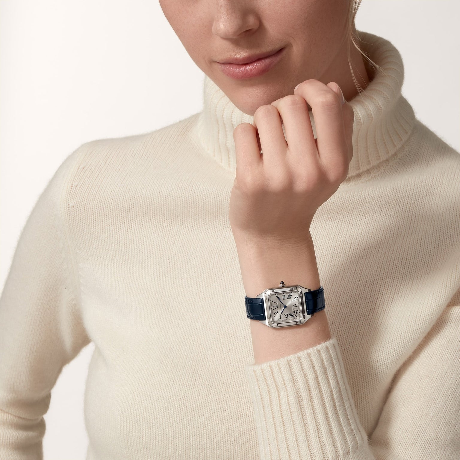 A person in a cream turtleneck sweater wears a Cartier Santos-Dumont watch with a square face and blue strap on their left wrist.