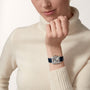 A person in a cream turtleneck sweater wears a Cartier Santos-Dumont watch with a square face and blue strap on their left wrist.