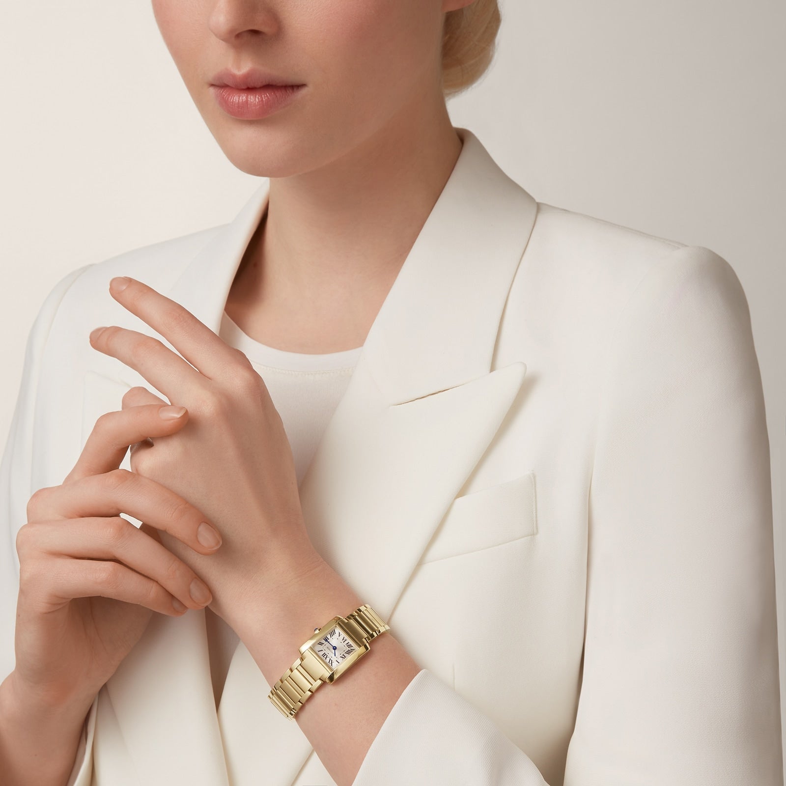 A woman in a white blazer and top wears the Cartier Tank Française on her left wrist, showcasing its yellow gold case with her hands poised near her chest.