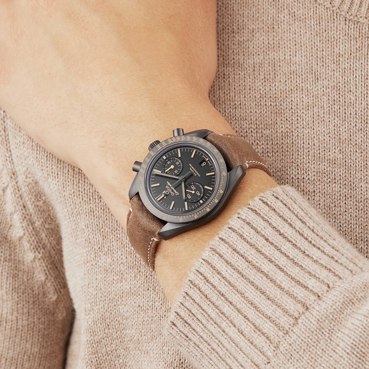 Someone in a brown sweater wears an OMEGA Speedmaster Dark Side Of The Moon on their left wrist, featuring a dark face, three subdials, and a brown leather strap.