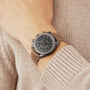Someone in a brown sweater wears an OMEGA Speedmaster Dark Side Of The Moon on their left wrist, featuring a dark face, three subdials, and a brown leather strap.