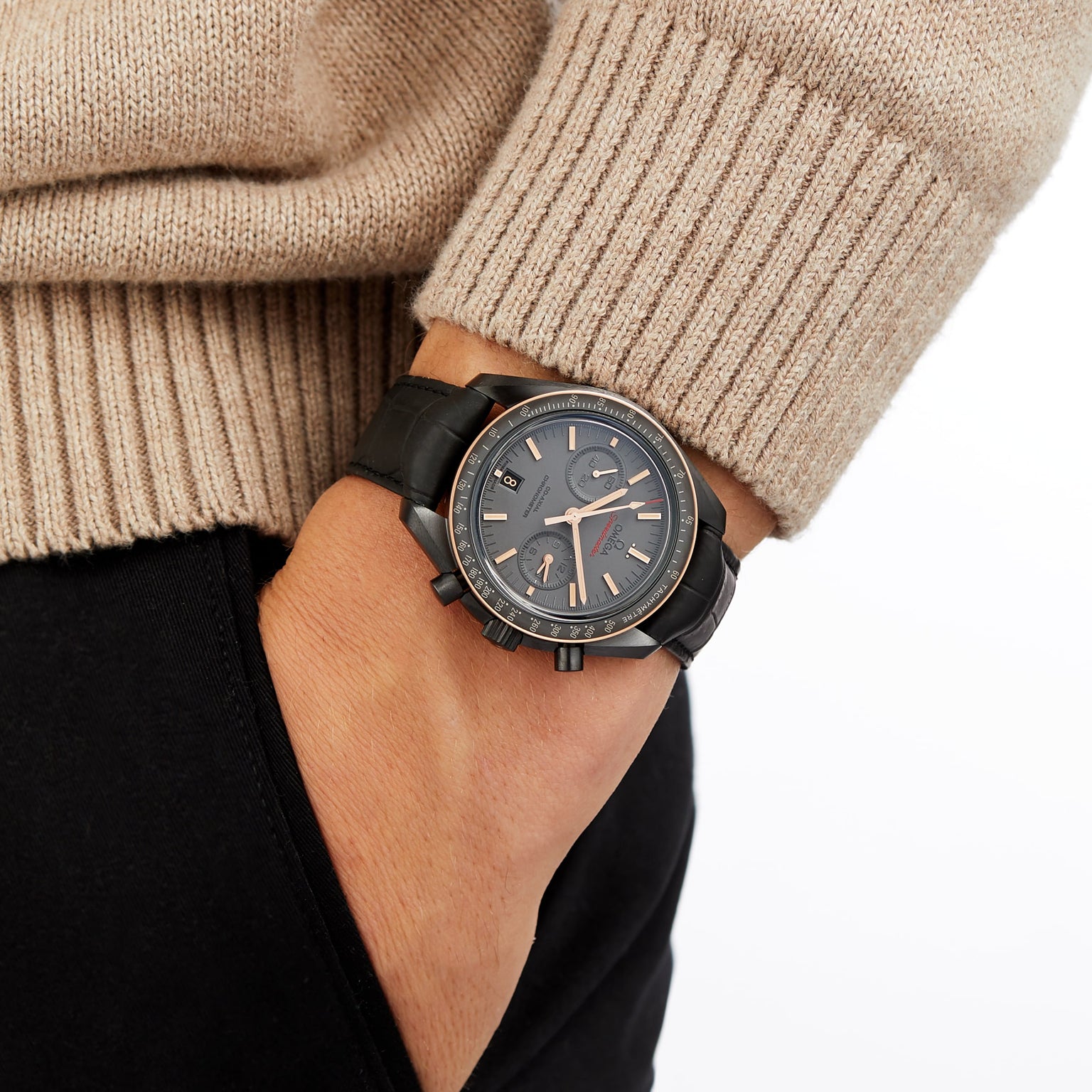 A person in a beige sweater and black pants has their left hand in their pocket, showing an OMEGA Speedmaster Dark Side Of The Moon Sedna Black chronograph with a round dark face and multiple dials.