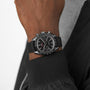 Wearing an OMEGA Speedmaster Dark Side of The Moon Ceramic chronograph with a textured strap and a black jacket, against a plain light background.