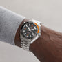 A person wearing an OMEGA Seamaster Planet Ocean 600M with a black and orange dial, black ceramic bezel, stainless steel band, and a gray long-sleeve shirt.