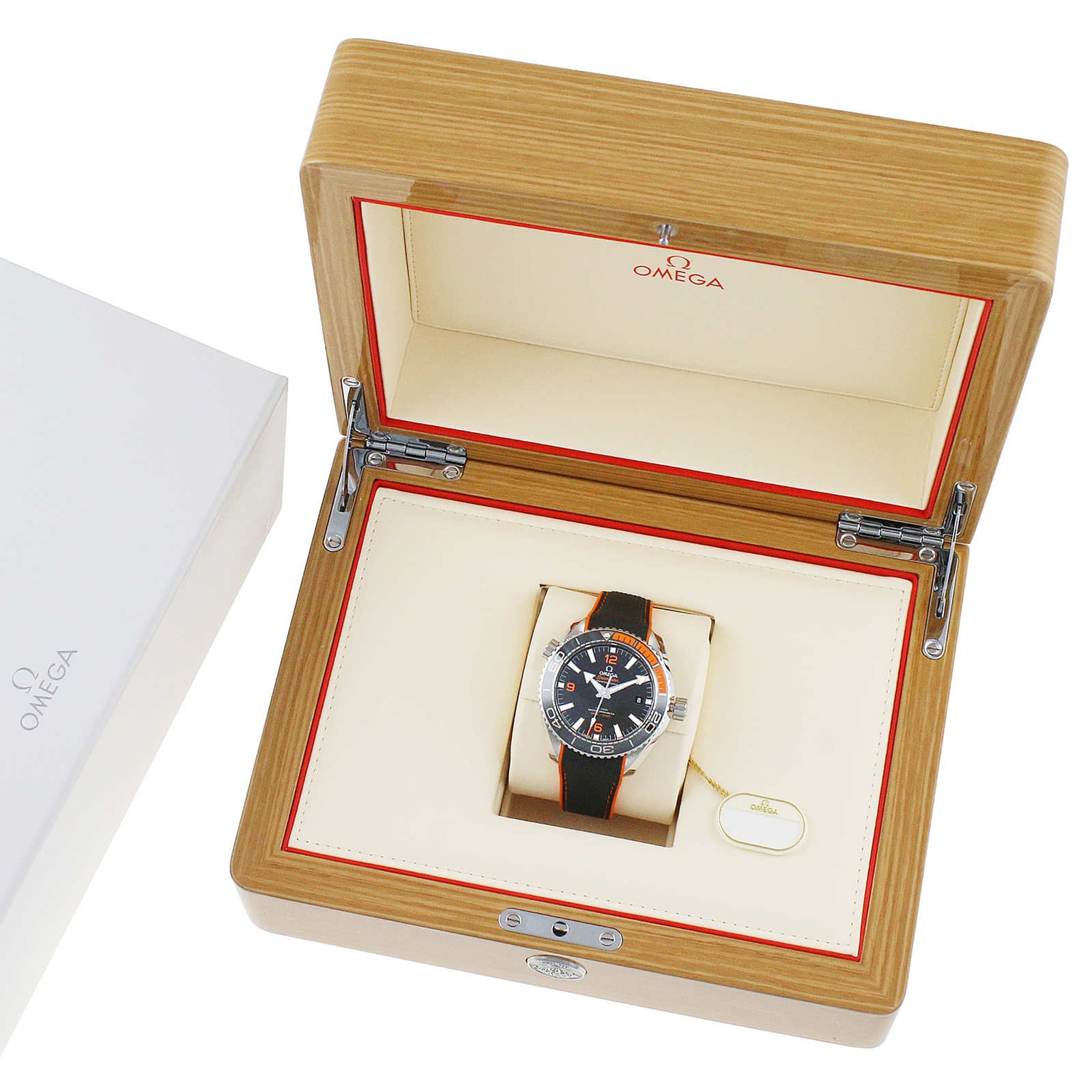 An OMEGA Seamaster Planet Ocean 600M dive watch with a black and orange strap is displayed in an open wooden OMEGA box with cream and red interior, next to a white OMEGA outer box.