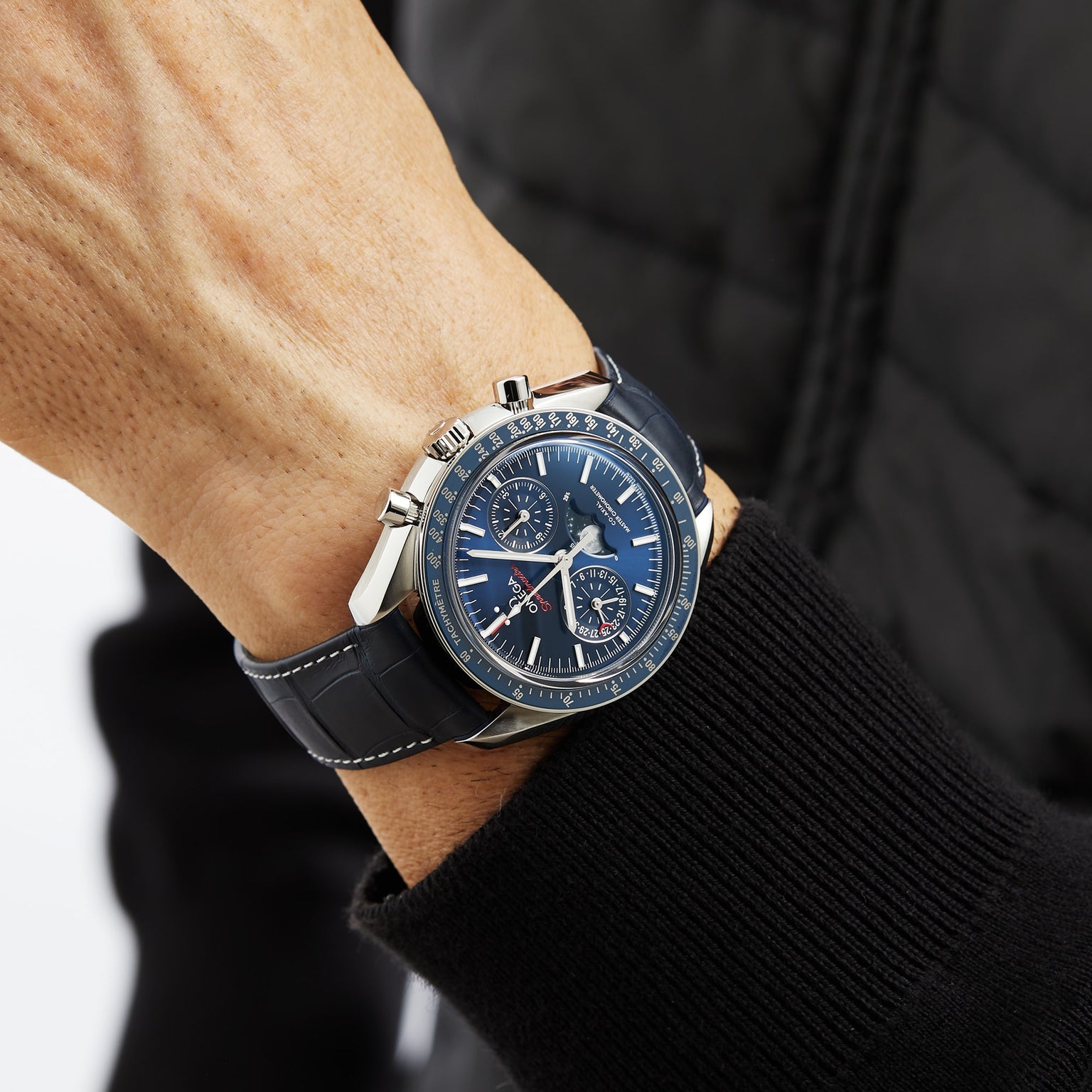 A close-up of a person in a black quilted jacket wearing an OMEGA Speedmaster Moonphase with a blue chronograph dial and leather strap on their left wrist.