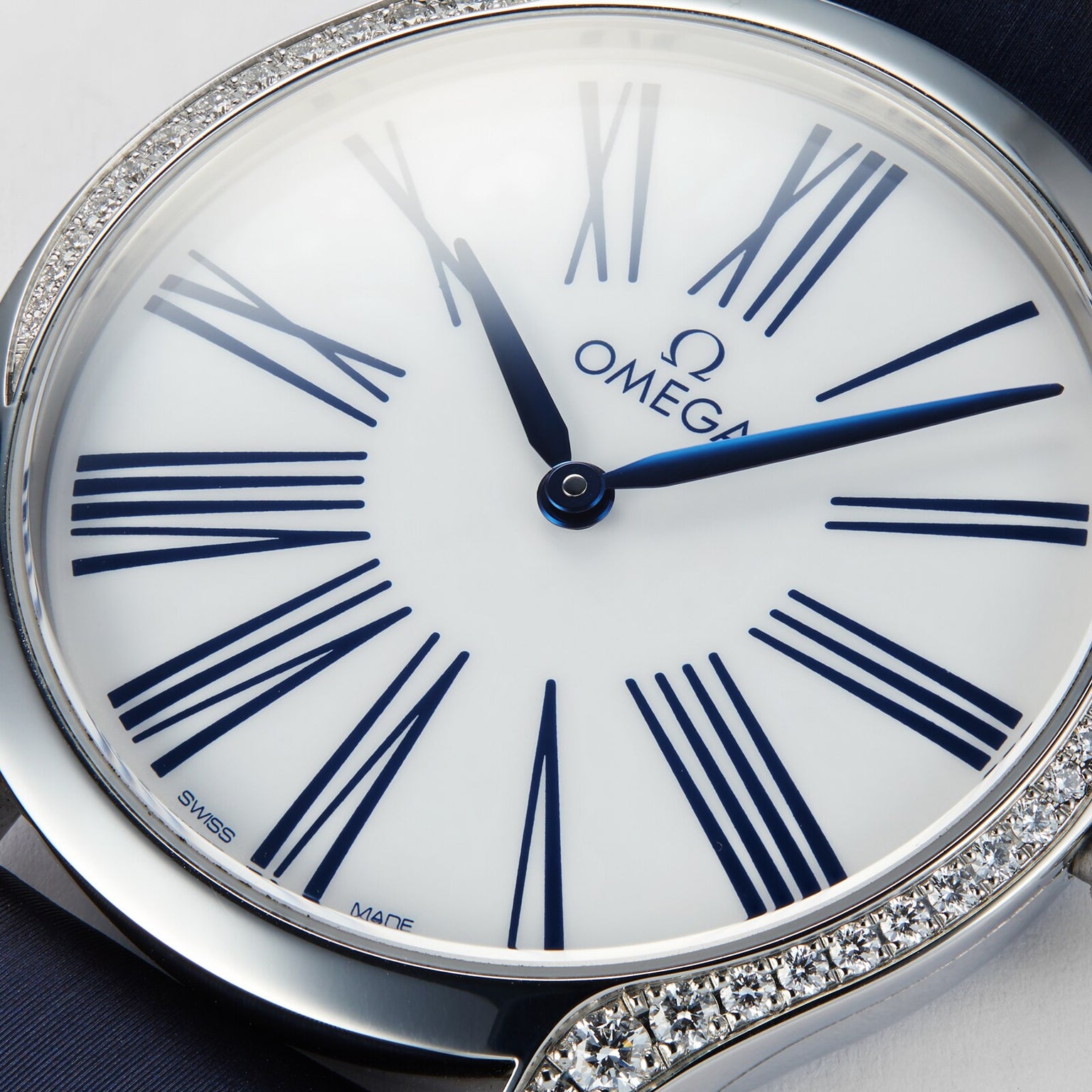 A close-up of the OMEGA De Ville Tresor 36 ladies watch showcasing blue Roman numerals and hands, a white dial, and a diamond-set bezel. The luxury timepiece displays the time at about 10:10.