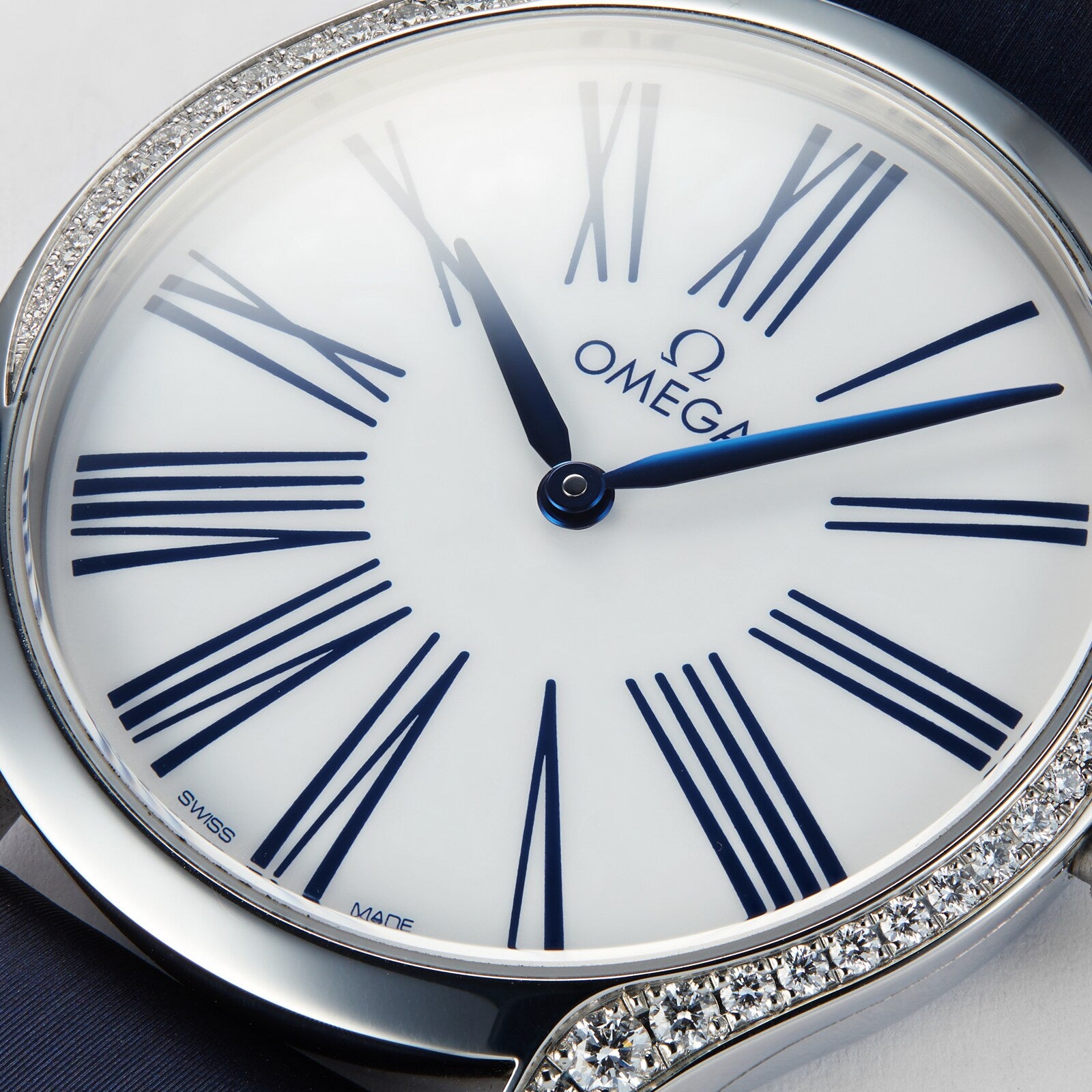 A close-up of the OMEGA De Ville Tresor 36 ladies watch showcasing blue Roman numerals and hands, a white dial, and a diamond-set bezel. The luxury timepiece displays the time at about 10:10.