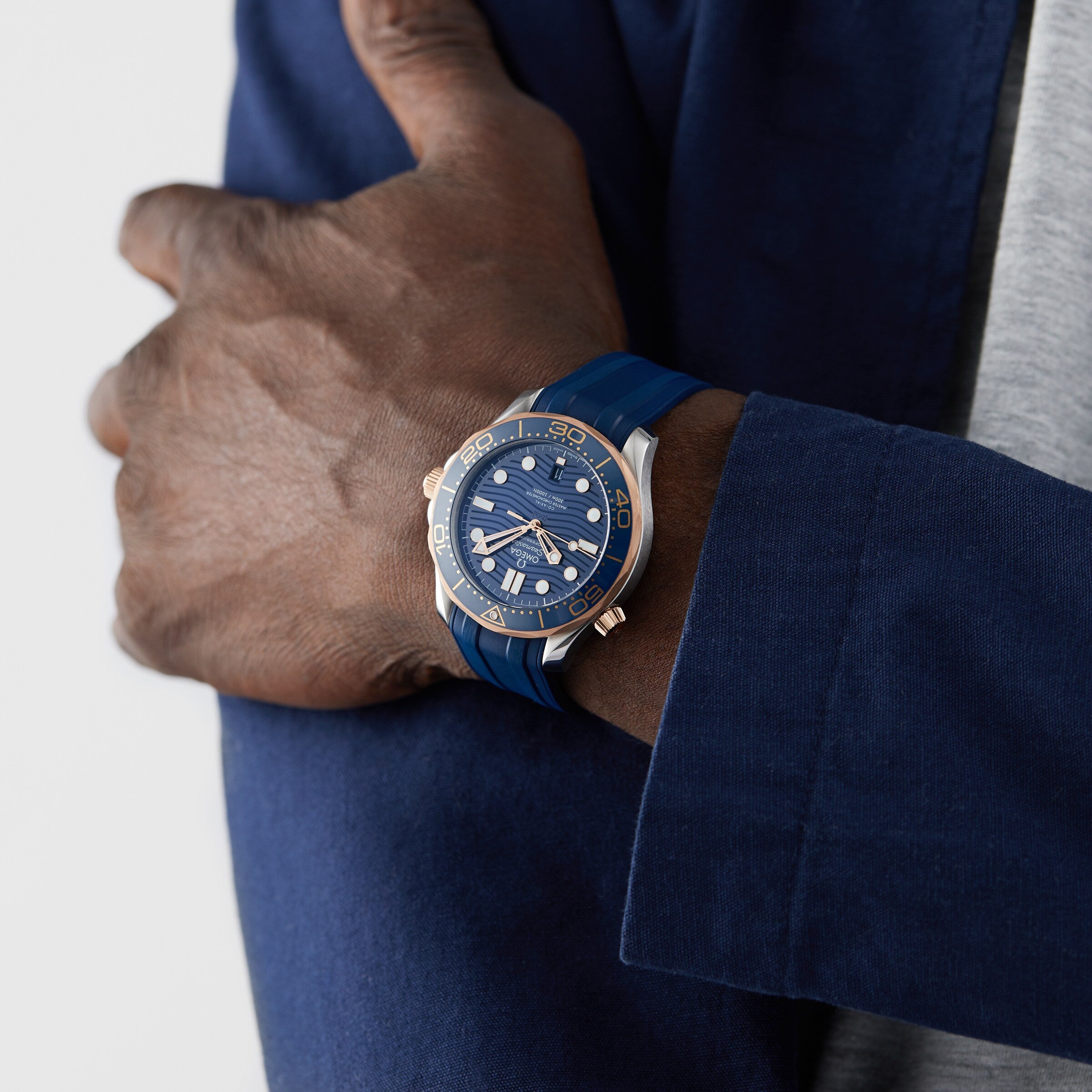 A person in a blue jacket and gray shirt stands with arms crossed, wearing an OMEGA Seamaster Diver 300M with a blue strap, round face, and powered by the OMEGA Co-Axial calibre 8800.