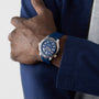 A person in a blue jacket and gray shirt stands with arms crossed, wearing an OMEGA Seamaster Diver 300M with a blue strap, round face, and powered by the OMEGA Co-Axial calibre 8800.